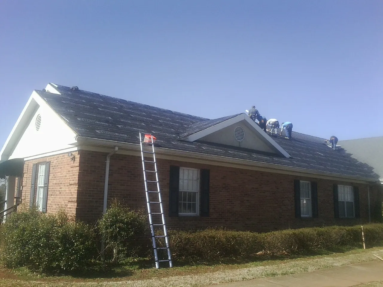 Crew tearing shingles off a brick ranch roof with a ladder set — Emergency Roof Repair work by our crew in Union Grove